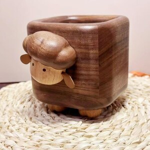 May include: A wooden planter shaped like a cube with a sheep design. The sheep's head and ears are made of light-colored wood, while the cube is a dark brown. The planter sits on small, rounded feet.
