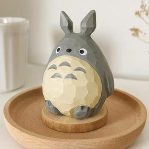 May include: A carved wooden figurine of Totoro, a character from the animated film My Neighbor Totoro. The figure is gray with a cream-colored belly and sits on a small wooden base. It is displayed on a wooden tray.