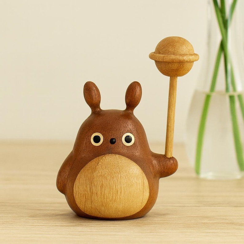 May include: A wooden figurine of a cartoon character with a brown body, light brown belly, and small ears. The character holds a wooden lollipop. The eyes are gold-colored. The figurine is on a wooden surface.