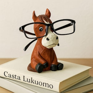 Könnte beinhalten: Eine geschnitzte Holzpferdefigur in Braun und Weiß, mit einer schwarzen Brille. Das Pferd sitzt auf einem Stapel Bücher, mit dem Titel "Casta Lukuomo" sichtbar. Ein dekoratives Element.