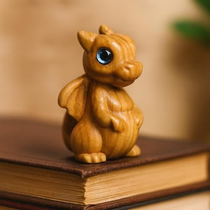 Könnte beinhalten: Eine Holzdrachenfigur mit blauen Augen und kleinen Flügeln. Der Drache ist aus hellfarbenem Holz geschnitzt und hat einen freundlichen Ausdruck. Die Figur sitzt auf einem braunen Buch.