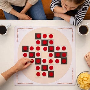 May include: A board game with red and black circular pieces arranged on a white and beige board. The board is surrounded by decorative arrows and lines. Two mugs of coffee and a bowl of chips are on the table. Several people are gathered around the table.
