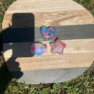 May include: Three colorful, handmade resin shapes: a heart, a fish, and a star. The heart is filled with colorful beads, the fish is blue and purple, and the star is pink. They are displayed on a wooden, striped serving board.