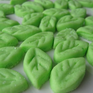 May include: A close-up of a collection of green leaf-shaped candies. The candies are arranged in a random pattern on a white surface.