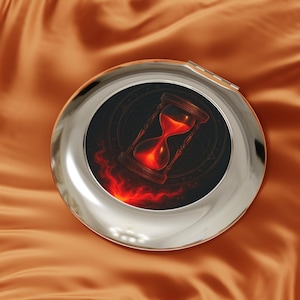 Könnte beinhalten: Ein runder, silberner Kompaktspiegel mit Klappdeckel. Die Spiegelfläche zeigt eine detaillierte Illustration einer roten Sanduhr mit Flammen, vor einem dunklen Hintergrund. Das Design hat ein mystisches oder Fantasy-Thema.