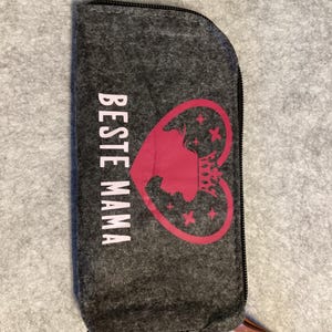 Könnte beinhalten: Eine dunkelgraue Filztasche mit Reißverschluss und dem vertikal aufgedruckten weißen Schriftzug BESTE MAMA auf der linken Seite. Rechts befindet sich ein rosa Herzumriss mit der rosa Silhouette einer gekrönten Person und kleinen rosa Sternen.
