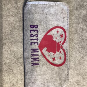 Könnte beinhalten: Ein hellgraues Filz-Etui mit Reißverschluss. Auf der Vorderseite ist der violette Schriftzug BESTE MAMA vertikal neben einem roten Herz-Motiv aufgedruckt, das eine Kronen-Silhouette und mehrere kleine vierzackige Sterne enthält.