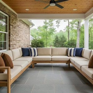 Può includere: Un ampio divano componibile da esterno a forma di U con cuscini beige e struttura in legno. Cuscini decorativi blu navy e marroni aggiungono contrasto. Il divano è su un patio in pietra, con vista sugli alberi e un muro di mattoni.