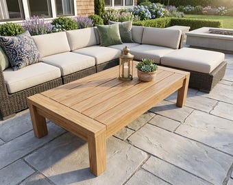 May include: A rectangular wooden coffee table in light brown with a slatted top, on a stone patio. A beige sectional sofa with woven brown sides is in the background. Decorative cushions and a lantern are on the table.