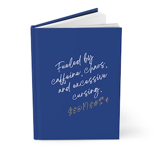 Op de afbeelding: Een hardcover notitieboek met een donkerblauwe kaft. De kaft heeft witte cursieve tekst: "Fueled by caffeine, chaos, and excessive cursing." Gouden symbolen staan eronder. Het notitieboek is open.