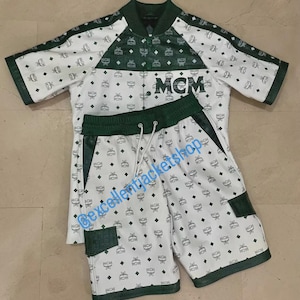 May include: White and green matching set featuring a short-sleeved jacket and shorts. The set is patterned with a repeating logo design. The jacket has the letters "MCM" on the front. The shorts have a drawstring waist.