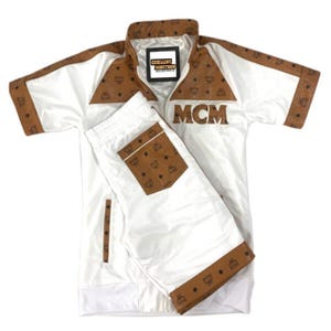 May include: White tracksuit set with brown accents and the MCM logo. The jacket has a collared neckline and the shorts have a drawstring waist. The brown accents feature a repeating pattern. The text "EXCELLENT JACKETSHOP" is visible on the jacket.