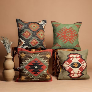May include: Four square throw pillows featuring woven geometric patterns in shades of red, green, brown, black, and cream. The pillows display intricate diamond and stepped motifs. A tall, light brown ceramic vase stands to the left of the pillows.