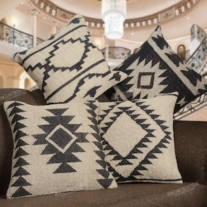 May include: Four square throw pillows with geometric patterns in black and beige. The pillows are arranged on a brown sofa, showcasing various tribal-inspired designs. The pillows add a decorative touch to the living space.
