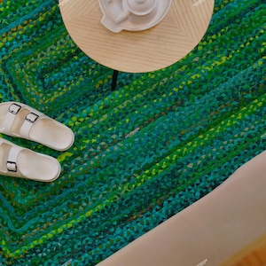 May include: An overhead shot of a green and blue braided rug with a pair of white sandals. The sandals have two black buckles. A small, round table and a decorative object are in the background.