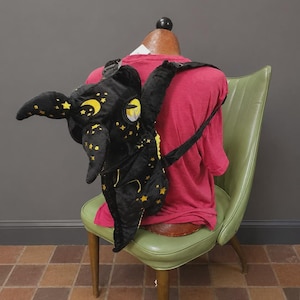 May include: A black plush backpack shaped like a creature with large pointed ears and wings. The fabric features embroidered yellow stars and crescent moons. It includes adjustable black straps and a single large eye detail on the side of the head.