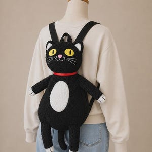 May include: A black cat-shaped backpack made of textured, fuzzy material. The cat features large yellow eyes, a pink nose, white inner ears, a white belly patch, and white paw tips. A red ribbon collar is around the neck, and black straps are attached.