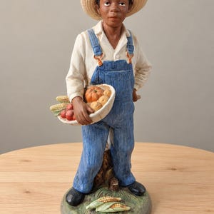 May include: A figurine of a person wearing a straw hat, a white collared shirt, and blue denim overalls. The figure holds a white basket filled with orange pumpkins and red apples, while two ears of corn lie on the green base at their feet.