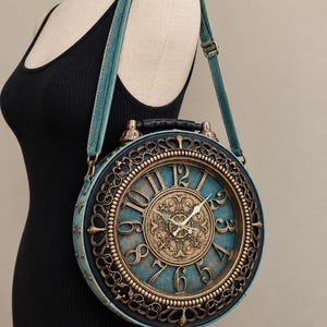 May include: A round shoulder bag designed to resemble an antique alarm clock. The bag features a teal blue face with gold-toned Arabic numerals, ornate filigree detailing, and a black handle on top. It includes a matching teal adjustable shoulder strap.