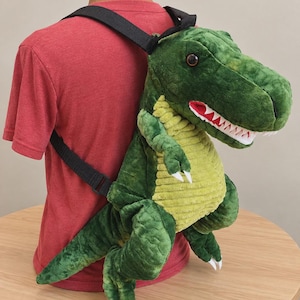 Könnte beinhalten: Ein Plüsch-Rucksack in Form eines Tyrannosaurus rex mit dunkelgrünem, strukturiertem Körper, einem hellgrünen Bauch in Kordoptik, weißen Filzzähnen und verstellbaren schwarzen Nylongurten. Der Dinosaurierkopf hat dunkelbraune Augen und ein offenes Maul.