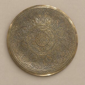 May include: A circular brass plate featuring intricate, deeply engraved patterns. The design consists of a central medallion surrounded by concentric bands of swirling, interconnected motifs and geometric borders across the surface.
