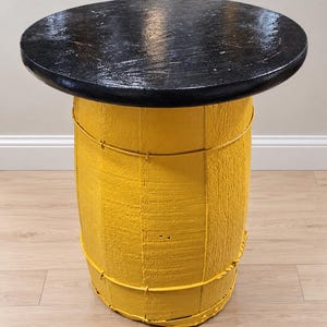 May include: A round table constructed from a repurposed wooden barrel. The base is painted a bright yellow and features horizontal metal bands. The top is a circular, flat wooden surface finished in a glossy, textured black paint.