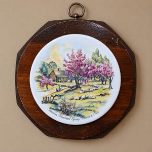 May include: A circular ceramic wall plaque mounted on a dark brown octagonal wooden frame with a metal hanging ring. The ceramic center features a colorful landscape painting of a house, pink flowering trees, and sheep, labeled American Homestead Spring Carrier & Ives.