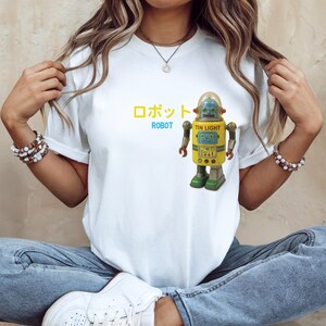 以下が含まれることがあります： ヴィンテージスタイルのロボットイラストが特徴の白いTシャツ。ロボットは黄色、青、緑色で、「ROBOT」の文字と日本語の文字が上にあります。「TIN LIGHT」の文字もロボットに印刷されています。