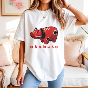 以下が含まれることがあります： 赤い赤べこのイラストがプリントされた白いTシャツ。黒いアクセントと赤い文字で「akabeko」と書かれています。赤べこは日本の伝統的なおもちゃです。カジュアルな半袖スタイルです。