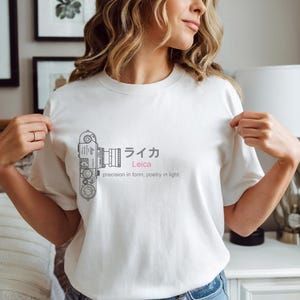 以下が含まれることがあります： ライカカメラの詳細な線画が特徴の白いTシャツ。デザインには日本語の文字とピンク色の英語の「Leica」、そして「precision in form, poetry in light.」というフレーズが含まれています。