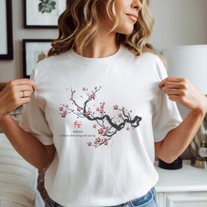 以下が含まれることがあります： 白いTシャツ。日本の桜の枝のデザイン。デザインには日本語の「桜」と「a flower that brings the spring」というフレーズが含まれています。枝は黒で、赤い花が咲いています。