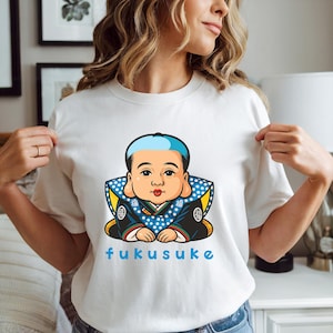 K&ouml;nnte beinhalten: Wei&szlig;es T-Shirt mit einer Cartoon-Illustration einer japanischen Puppe, FUKUSUKE, mit einem blau-wei&szlig; gepunkteten Kragen und blauem Text. Die Puppe hat eine blaue M&uuml;tze und tr&auml;gt eine dunkelblaue Robe mit goldenen Akzenten.