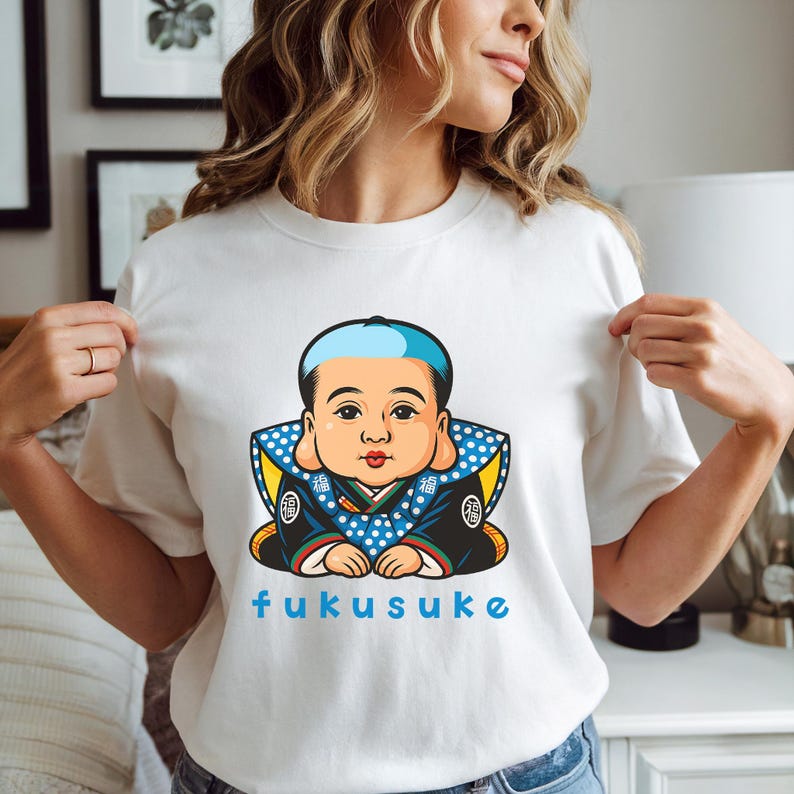 K&ouml;nnte beinhalten: Wei&szlig;es T-Shirt mit einer Cartoon-Illustration einer japanischen Puppe, FUKUSUKE, mit einem blau-wei&szlig; gepunkteten Kragen und blauem Text. Die Puppe hat eine blaue M&uuml;tze und tr&auml;gt eine dunkelblaue Robe mit goldenen Akzenten.