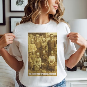 以下が含まれることがあります： ロボットのようなマスクをつけた家族のセピア色の画像がプリントされた白いTシャツ。「THE FAMILY THAT UPGRADED TOGETHER」というテキストが下部の長方形のボックスに表示されています。家族は建物と木の前に立っています。