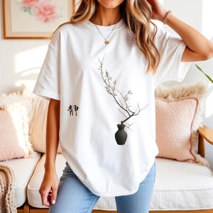 Könnte beinhalten: Weißes T-Shirt mit einem minimalistischen Design einer schwarzen Vase, die einen Zweig mit weißen Blüten hält. Das T-Shirt hat auch das japanische Zeichen für "Ruhe" aufgedruckt. Das T-Shirt ist aus einem weichen Material.