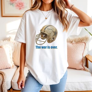 以下が含まれることがあります： 白のTシャツ。使い古されたアメフトのヘルメットのグラフィック、星、そして「Peace！」の文字がプリントされています。「The war is over」の文字が青色でプリントされています。カジュアルな普段着です。