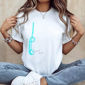 以下が含まれることがあります： 白いTシャツに、ターコイズブルーの電話のグラフィックと「HELLO？」の文字がプリントされています。デザインにはカールしたコードが含まれています。カジュアルな普段着です。