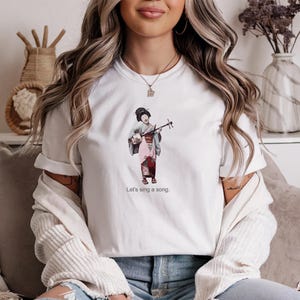 May include: White t-shirt featuring a watercolor illustration of a woman playing a traditional Japanese instrument. The image includes the text "Let's sing a song."