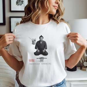 May include: White t-shirt with a black and white illustration of a Japanese man in traditional clothing, a bucket, and a sword. The text "切腹 Japanese HARAKIRI" is printed in red and black.