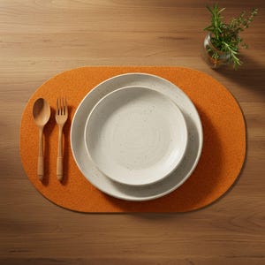 Könnte beinhalten: Draufsicht auf eine Tischdekoration. Ein orangefarbener, ovaler Tischset hält zwei gesprenkelte, weiße Teller. Ein Holzlöffel und eine Gabel befinden sich links. Eine kleine Glasvase mit Kräutern steht oben rechts.
