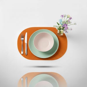 Puede incluir: Una mesa moderna con un mantel individual naranja quemado, un plato verde claro y un cuenco rosa. La cubertería de plata incluye un tenedor y un cuchillo. Un pequeño jarrón de flores coloridas añade un toque decorativo. El conjunto está sobre un fondo blanco.
