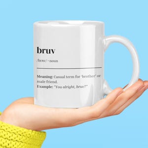 May include: A white mug featuring the word "bruv" with its pronunciation and definition. The mug is held in a hand against a light blue backdrop. The definition is a casual term for 'brother' or male friend.