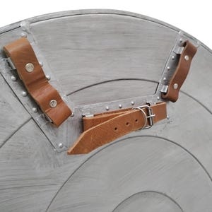 Pu&ograve; includere: Primo piano di uno scudo color argento con un design a cerchi concentrici. Lo scudo ha cinghie in pelle marrone e rivetti metallici per il fissaggio. Le cinghie sono fissate con fibbie e rivetti metallici.