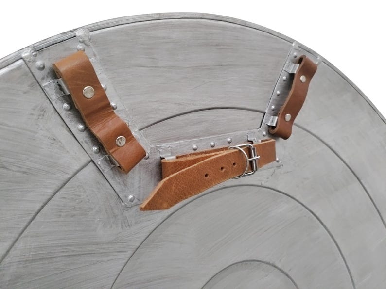 Pu&ograve; includere: Primo piano di uno scudo color argento con un design a cerchi concentrici. Lo scudo ha cinghie in pelle marrone e rivetti metallici per il fissaggio. Le cinghie sono fissate con fibbie e rivetti metallici.