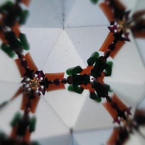 May include: Abstract kaleidoscope image with a geometric pattern. The design features orange, green, and burgundy elements arranged around a central point. The background is a light, geometric shape, creating a symmetrical and colorful visual.