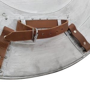 Pu&ograve; includere: Primo piano di uno scudo color argento con un cinturino e una fibbia in pelle marrone. Lo scudo ha un design circolare con rivetti e un aspetto usurato. Il cinturino in pelle &egrave; fissato con rivetti.