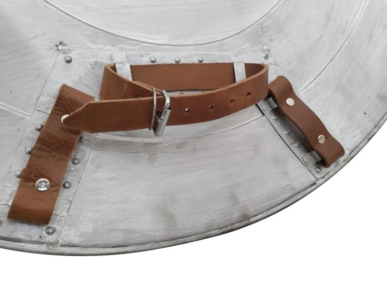 Pu&ograve; includere: Primo piano di uno scudo color argento con un cinturino e una fibbia in pelle marrone. Lo scudo ha un design circolare con rivetti e un aspetto usurato. Il cinturino in pelle &egrave; fissato con rivetti.