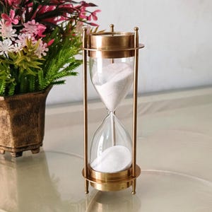 Pode incluir: Uma ampulheta de latão com câmaras de vidro transparente preenchidas com areia branca. A ampulheta tem um design clássico com uma parte superior e inferior circulares e hastes de suporte verticais. Um arranjo floral decorativo está no fundo.