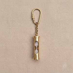 May include: A gold-toned metal keyring featuring a small, cylindrical hourglass pendant. The hourglass is encased in a polished brass-coloured frame with a matching chain and a teardrop-shaped ring at the top for attachment to keys or bags.