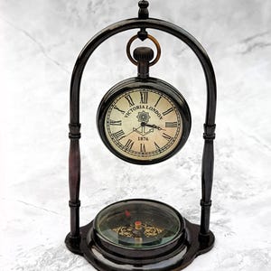 May include: A vintage desk clock featuring a compass. The clock face displays "Victoria London 1876" with Roman numerals. The clock is held by a dark metal frame with wooden detailing. The compass is at the base, below the clock.
